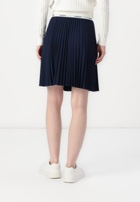 Navy pleated skirt with an elastic waistband featuring a white logo. Worn with white sneakers, showcasing a simple, clean design.