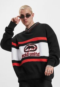 Black oversized sweatshirt with red and white horizontal stripes, featuring a graphic logo. Ribbed neckline and cuffs. Casual fit.