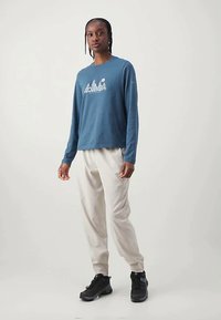 Cămașă albastră cu mâneci lungi, cu grafică „Columbia”, asortată cu pantaloni de culoare deschisă și pantofi sport negri, pe un fundal neutru.