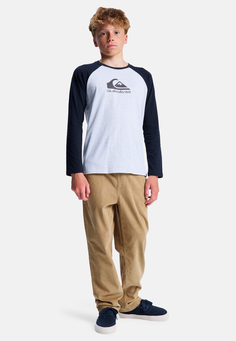 Grijze en marineblauwe langekleurige shirt met een logo, gecombineerd met tan corduroybroek en marineblauwe schoenen. Model staat tegen een eenvoudige achtergrond.