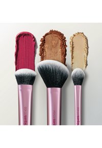 Trois pinceaux de maquillage à manche rose avec des poils noirs et blancs, chacun estompant différentes couleurs de maquillage : rose profond, bronze métallique et beige clair.