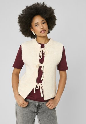 Femme avec une coiffure afro naturelle portant un gilet en tricot crème noué avec des nœuds par-dessus une chemise bordeaux et un jean gris sur un fond uni.