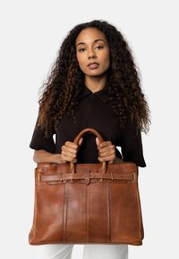 Una donna con lunghi capelli ricci indossa una maglia scura e pantaloni bianchi, tenendo una grande borsa in pelle marrone con doppi manici davanti a uno sfondo bianco.