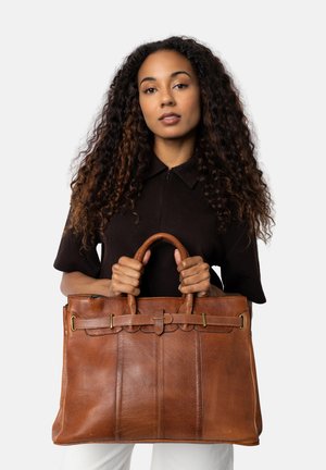 Una donna con lunghi capelli ricci indossa una maglia scura e pantaloni bianchi, tenendo una grande borsa in pelle marrone con doppi manici davanti a uno sfondo bianco.