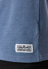 Blue textured fabric short-sleeve shirt with a woven pattern, featuring a white label on the hem with "DENIM FACTORY" printed on it.