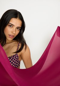 Lyse magenta gjennomsiktig stoff drapert i hånden, kombinert med en mønstret ermeløs topp med mørkerøde, hvite og blå former.