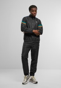 Chándal deportivo negro con chaqueta con cremallera que presenta franjas turquesas y naranjas, combinado con pantalones a juego y zapatillas de color claro.