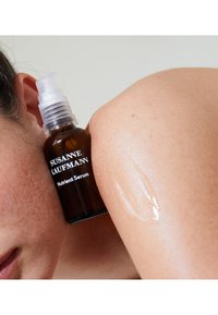 Brązowa szklana butelka z przezroczystym pompką, oznaczona etykietą "Susanne Kaufmann Nutrient Serum." Krople serum na jasnej skórze o gładkiej fakturze.