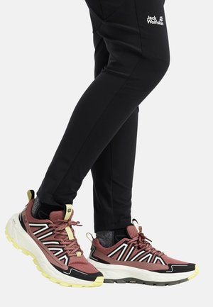 Benen in zwarte Jack Wolfskin broeken met bordeauxrode, zwarte en witte trailrunningschoenen met gele zolen en grijze sokken op een lichte achtergrond.