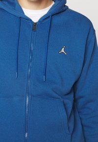 Homme portant un sweat à capuche bleu zippé avec cordons et un petit logo blanc Jumpman sur la poitrine gauche, par-dessus une chemise blanche.