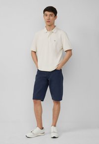 Polo crème à manches courtes, shorts navy et baskets blanches. Le polo présente un petit logo sur la poitrine. Les shorts sont à mi-genoux.