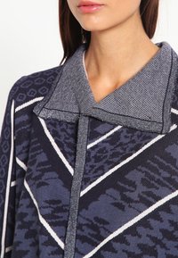 Mörkblå mönstrad jacka med stor krage, geometriska mönster och kontrasterande sömmar längs kanterna. Mjuk textur och avslappnad passform.