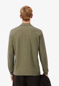 Camisa polo de manga larga en verde oliva, con un cuello plano y tejido texturizado. El modelo sostiene una prenda negra en una mano.