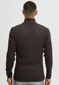 Strikket mørkebrun sweater med høj krave, rillemønster, lange ærmer og tætsiddende silhuet, som viser detaljerede stikmønstre.