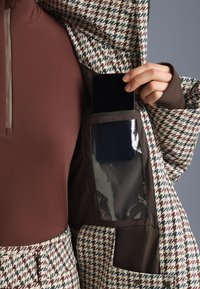 Jakna s uzorkom u houndstooth dizajnu u smešanoj, krem i plavoj boji. Džep s prozirnim poklopcem drži pametni telefon, a unutrašnjost je smeša boje smešane.