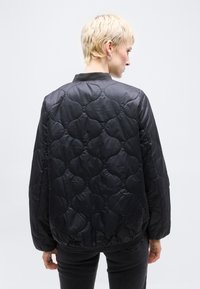 Άτομο με κοντά ξανθά μαλλιά που φοράει μαύρο μπουφάν με κουκούλα και ραφή σε σχήμα τριφυλλιού, γυρισμένο προς τα πίσω μπροστά σε απλό φόντο.