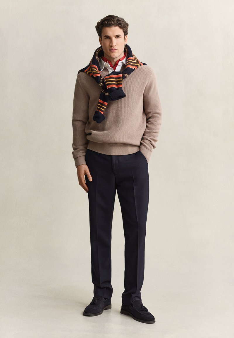 Man wearing beige sweater with striped scarf over shoulders, red shirt, dark trousers, and black shoes standing against plain background.