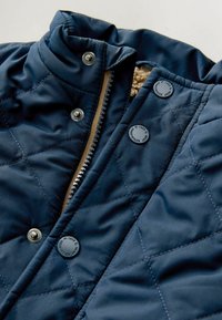 Navy quilted jacket with a high collar. Features silver hardware, a zipper, and buttons. Interior lined with textured beige fabric.