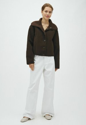 Femme portant un manteau boutonné marron foncé avec un col en peau lainée, un pantalon blanc à jambes larges et des chaussures plates beiges, debout devant un fond uni.