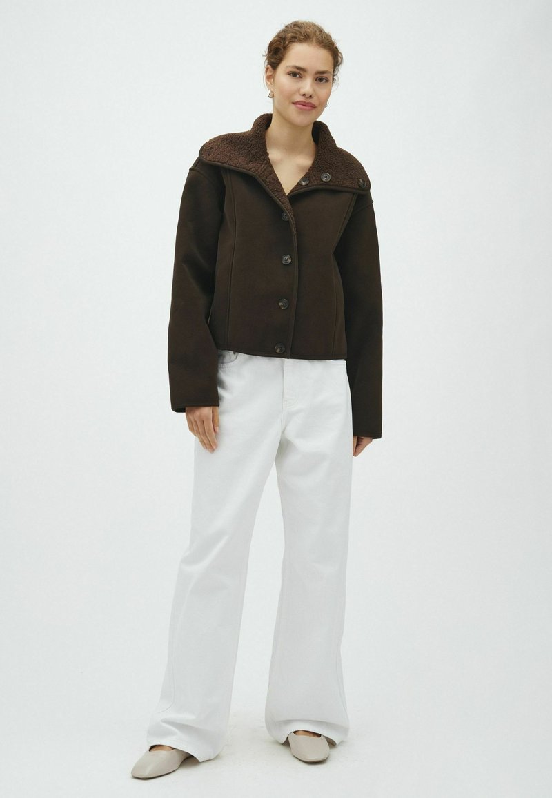 Femme portant un manteau boutonné marron foncé avec un col en peau lainée, un pantalon blanc à jambes larges et des chaussures plates beiges, debout devant un fond uni.