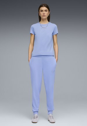 Junge Frau steht mit den Händen in den Taschen, trägt ein passendes hellblaues T-Shirt und Sweatpants mit weißen Turnschuhen vor grauem Hintergrund.