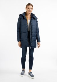 Navy gewatteerde jas met een ritssluiting aan de voorkant, grote capuchon en twee zijzakken. Draag deze over een wit shirt met donkere spijkerbroek en blauwe sneakers.