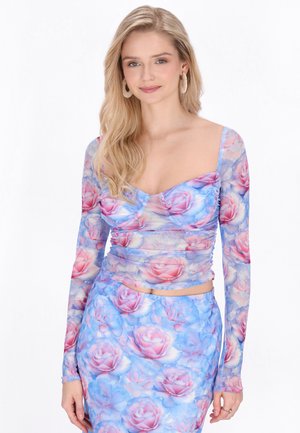 Młoda kobieta o długich blond włosach, ubrana w niebiesko-różową kwiatową bluzkę z długim rękawem i dopasowaną spódnicę, na białym tle.