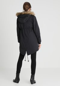 Svart parka med hette fôret med falsk pels, innsnevret midje og snører i kanten. Glatt stoff med matt tekstur og lange ermer.
