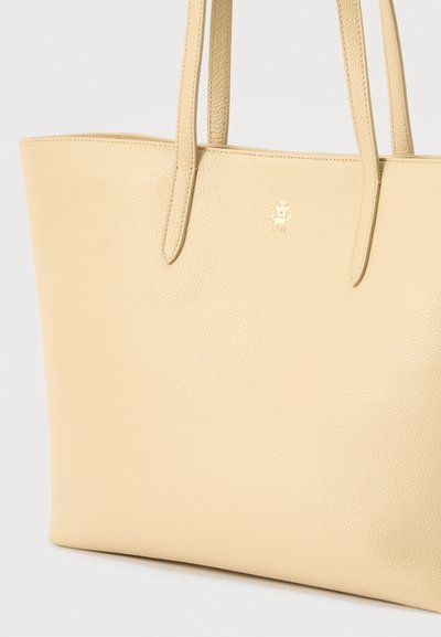 Μπεζ δερμάτινη τσάντα ώμου με υφασμάτινη υφή, διαθέτει δύο μακριές λαβές και χρυσό λογότυπο μπροστά. Απλή, ορθογώνια μορφή.