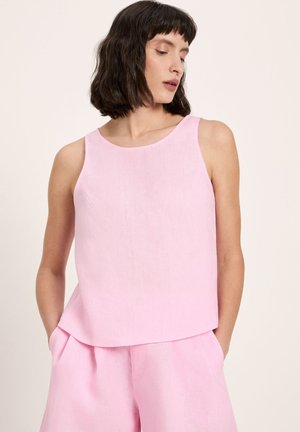 Frau mit dunklem Bob-Haar, die ein ärmelloses hellrosa Oberteil und passende weite Hose trägt, Hände in den Taschen, schaut nach rechts unten auf beigem Hintergrund.