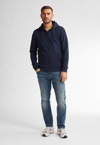 Navy zip-up hoodie with a hood and side pockets, paired with light blue jeans and white sneakers. Smooth fabric with no visible patterns.