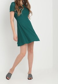 Tyrkysové pletené šaty s krátkými rukávy, rozšířenou sukní a texturovaným designem. Černé skluzavkové sandály s perforovaným vzorem.