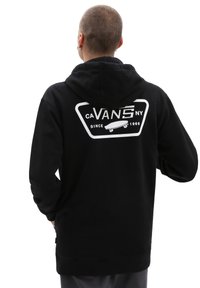 Hanorac negru cu un grafic mare alb pe spate care prezintă "VANS" și o placă de skateboard. Material moale, texturat, cu un buzunar în față.