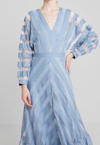 Ljusblå klänning med djup V-ringning, som har genomskinliga randiga ärmar och en midjebälte. Kjolen har en flödig design med en volangkant.