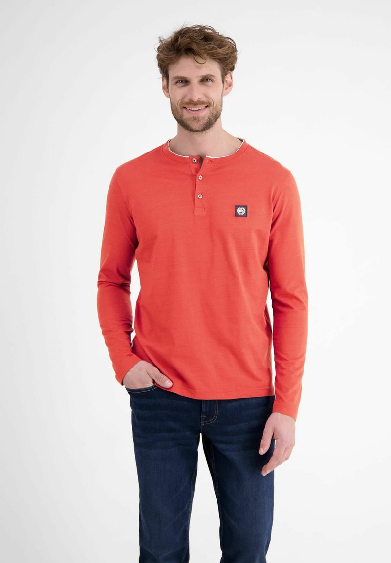 Langärmliges, orangefarbenes Henley-Shirt mit einem runden Halsausschnitt, einer Drei-Knopf-Leiste und einem kleinen Logo auf der linken Brust. Getragen mit dunklen Jeans.