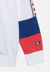 Manica di felpa bianca con strisce blu e rosse, polsini a coste e un piccolo logo ricamato. Texture liscia con colori a contrasto.