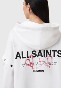 Sudadera blanca con un gran logotipo negro "ALLSAINTS" en la parte posterior, acompañado de gráficos de corazones rosas y texto japonés debajo. Material de algodón, diseño informal.