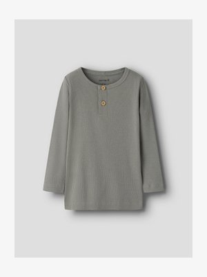 Ilgas rankovėmis, pilkas, riboto audinio marškinėlis mažyliui su apvalia iškirpte ir dviem rudos spalvos sagtelėmis priekyje ant apkausto, išdėliotas ant pilkos spalvos paviršiaus.