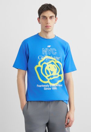 Jonge man draagt een felblauw NYC City park tour T-shirt met een grote gele roosafbeelding en grijze broek tegen een effen achtergrond.