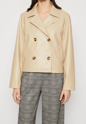 Veste beige courte à double boutonnage avec larges revers et grands boutons marron, associée à un pantalon taille haute à carreaux gris.