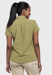 Olive green short-sleeve shirt with a structured collar, back pleat detail, and a rounded hem. Worn with light beige pants featuring an accent logo.