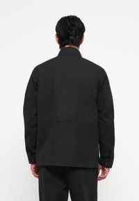 Black jacket made of smooth fabric, featuring a straight cut, long sleeves, and buttoned cuffs. Minimalist design with no visible patterns.