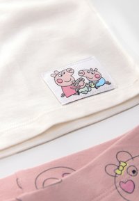 Beige cotton top featuring a logo patch with cartoon pigs. Pink bottom with pig face print, highlighted by yellow accents. Soft texture.