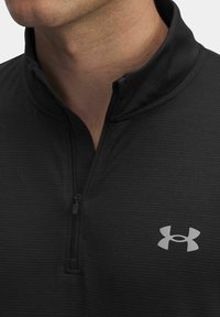 Man in zwart quarter-zip shirt van Under Armour, met de rits gedeeltelijk open, zichtbaar nek en onder gezicht tegen een effen achtergrond.