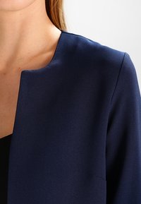 Marinblå topp med rundad halsringning, tillverkad av ett slätt tyg. Har en öppen framsida och en minimalistisk design utan synliga stängningar eller detaljer.