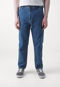 Jeans de denim azul con corte recto, cierre de cremallera, diseño de cinco bolsillos y ligero desgaste para un aspecto usado. Combinados con zapatillas grises.
