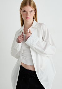 Jonge vrouw met lang blond haar, grote oorringen, een wit oversized button-up shirt, een wit geribbeld cropped top en zwarte broek.
