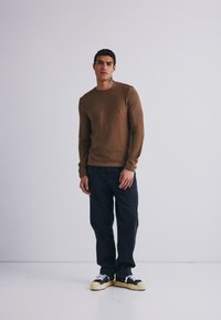 Suéter marrón de punto con escote redondo, combinado con jeans de mezclilla oscuros y zapatillas negras con acentos blancos y suelas color crema.