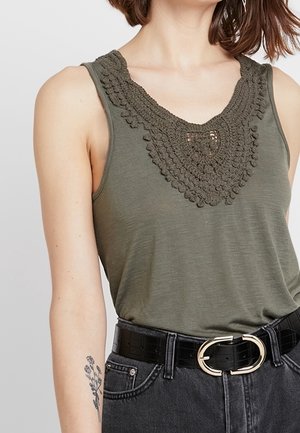 Kvinde iført olivengrøn ærmeløs top med hæklet udskæring, sort bælte med guldlås og mørke jeans, der viser en armtatovering.