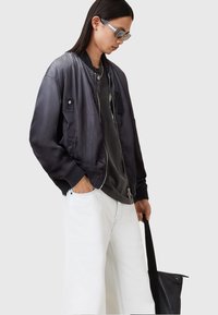 Bomber jacket dalam satin abu-abu gelap, penutupan ritsleting, manset bergaris, dan dua saku depan. Dipakai di atas kaos hitam dan celana putih; memegang tas tangan.